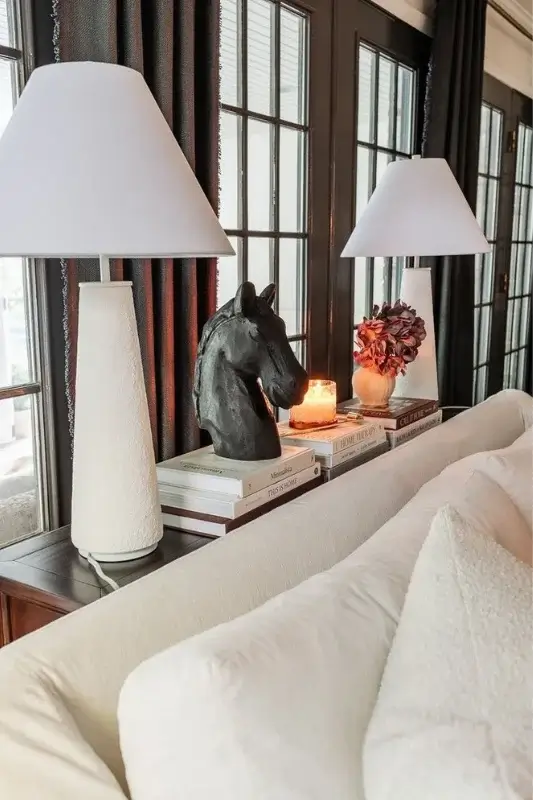 Sofa table behind couch styled with matching white lamps, stacked books, a black horse bust sculpture, and candles for symmetrical console table decor