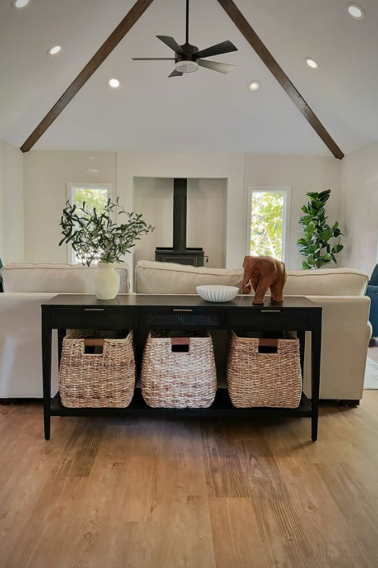 Rustic wood console table placed behind a soft gray sofa, styled with stacked books, a woven basket, ceramic vases, and natural greenery. The setup is anchored by a cozy stone fireplace and exposed wood beams for a warm, inviting living room feel.