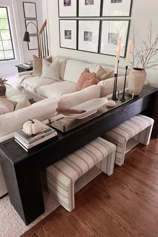 Dark wood console table behind a sectional sofa styled with candles, neutral decor, stacked books, and upholstered stools tucked underneath for functional sofa table seating