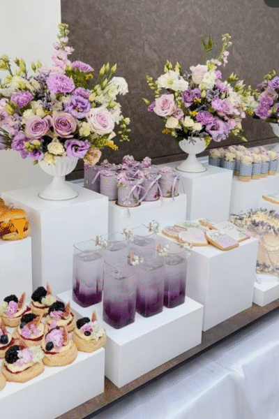 Lavender-themed graduation dessert table with purple floral arrangements, violet drinks, decorated cookies, cupcakes, and party favors displayed on white risers.