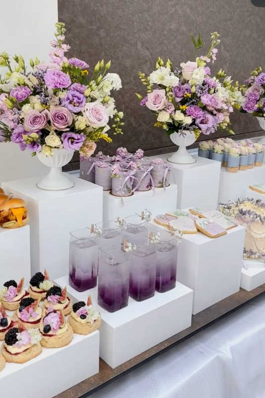 Lavender-themed graduation dessert table with purple floral arrangements, violet drinks, decorated cookies, cupcakes, and party favors displayed on white risers.