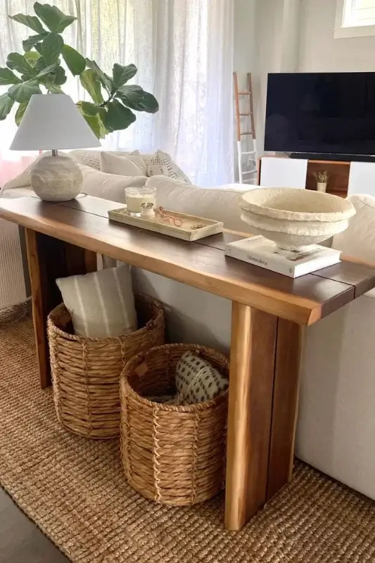 Live edge wood console table behind couch styled with woven baskets, stacked books, ceramic decor, and table lamp for warm modern living room styling