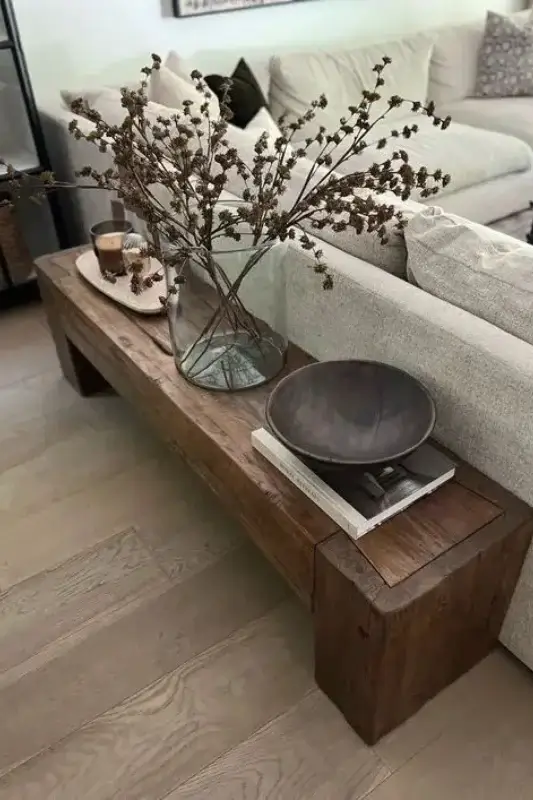 Low rustic wooden bench behind sofa styled with glass vase, dried branches, books, and decorative bowl for minimalist behind the couch decor