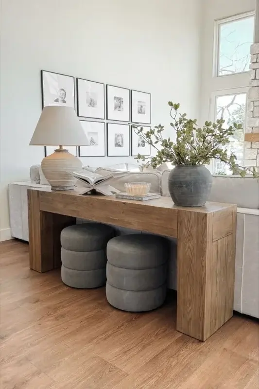 Modern wood console table behind sofa with round upholstered stools, table lamp, books, and greenery for functional and stylish living room decor