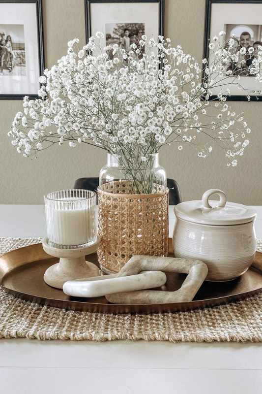 Elegant neutral centerpiece featuring a clear glass vase with baby's breath flowers inside a woven rattan sleeve, paired with a ribbed glass candle, a ceramic lidded jar, and modern sculptural links, all arranged on a round bronze tray over a woven jute table runner.