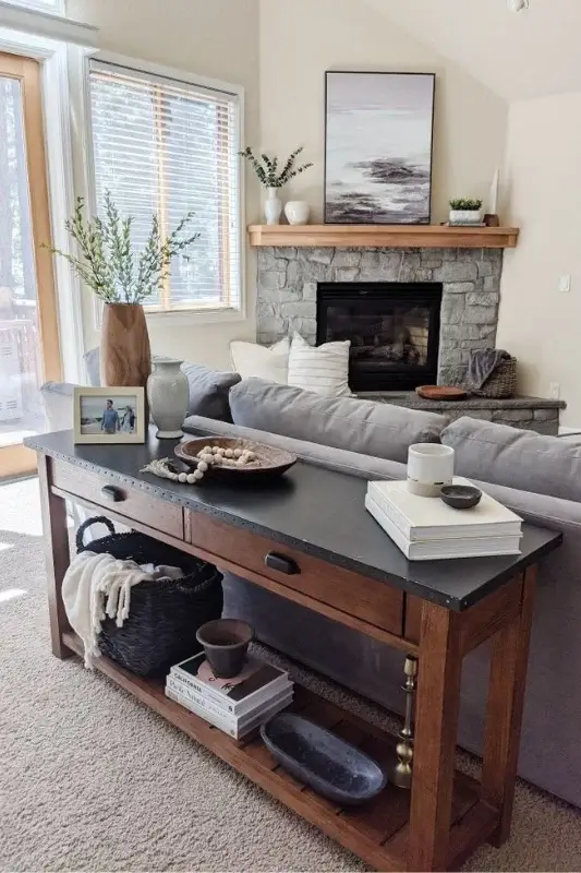 Rustic wood console table behind a gray sofa styled with books, basket storage, neutral decor, and a fireplace backdrop for cozy living room styling