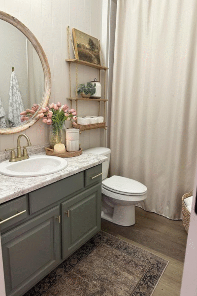 Vintage farmhouse bathroom featuring a round wood-framed mirror, sage green vanity with brass fixtures, a woven tray with candles and pink tulips, and a rope-hung wall shelf above the toilet displaying artwork, greenery, and paper goods.