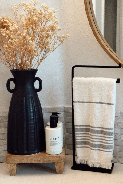 Chic farmhouse-style bathroom decor featuring a tall matte black vase with dried florals on a small wooden riser, a bottle of ELEMIS hand and body wash, and a white and gray striped towel on a minimalist black metal stand. A round wood-framed mirror adds warmth and balance to the vignette.