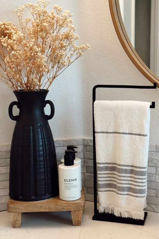 Chic farmhouse-style bathroom decor featuring a tall matte black vase with dried florals on a small wooden riser, a bottle of ELEMIS hand and body wash, and a white and gray striped towel on a minimalist black metal stand. A round wood-framed mirror adds warmth and balance to the vignette.