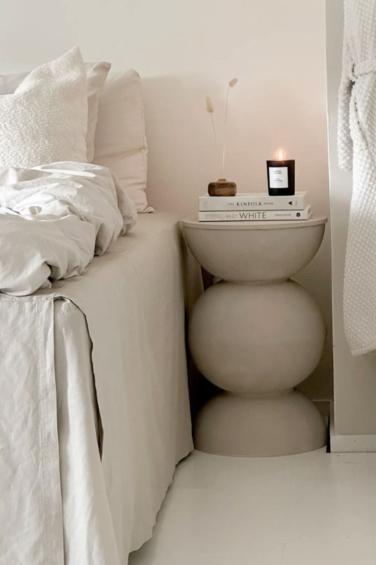 Neutral sculptural bedside table styled with books, candle, and dried flowers in a minimalist bedroom setting.