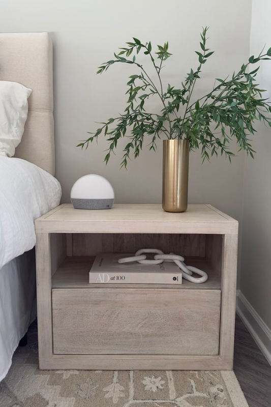 Light wood nightstand with brass vase, modern bedside lamp, and coffee table books in a clean, neutral bedroom setup.