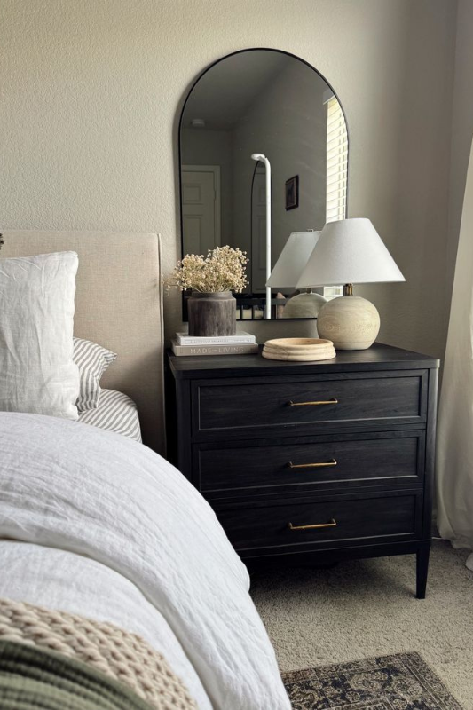 Matte black nightstand with mirror, ceramic lamp, stacked books, and soft accessories styled in a cozy neutral bedroom.