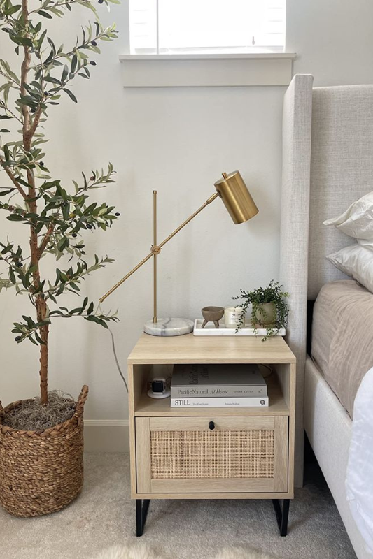 Light wood rattan nightstand with modern brass lamp, greenery, tray accessories, and books in a serene neutral bedroom.