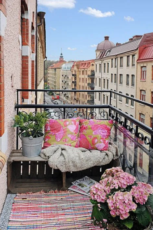 Tiny city balcony with bench seating, throw pillows, and flowers overlooking urban buildings