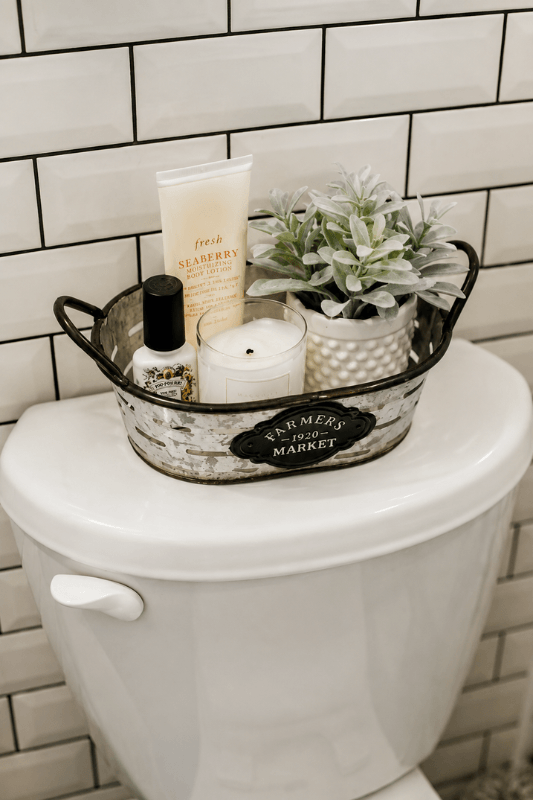 Farmhouse bathroom toilet tank decor with galvanized tray, candle, toiletries, and small potted greenery against white subway tile