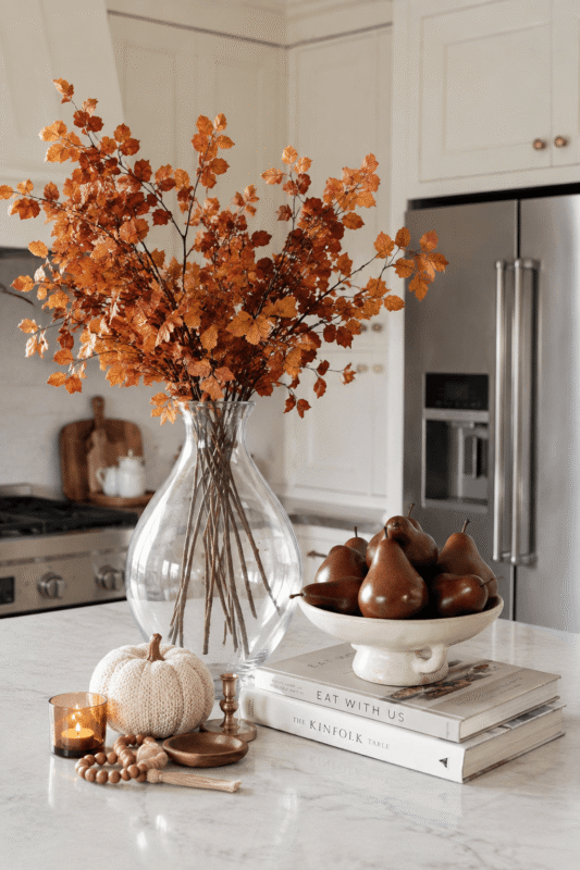 Fall kitchen centerpiece with brown pears, orange branches, knitted pumpkin, candle, and neutral cookbooks